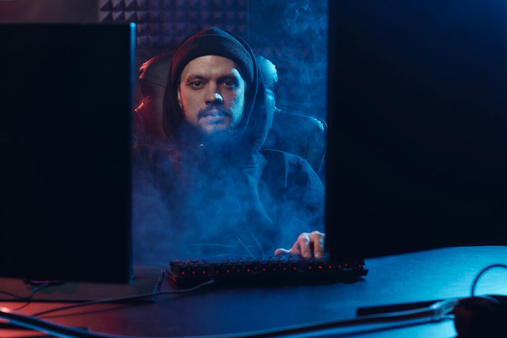 Dramatic scene of an adult man in a hoodie using a computer, surrounded by smoke in a dimly lit room.